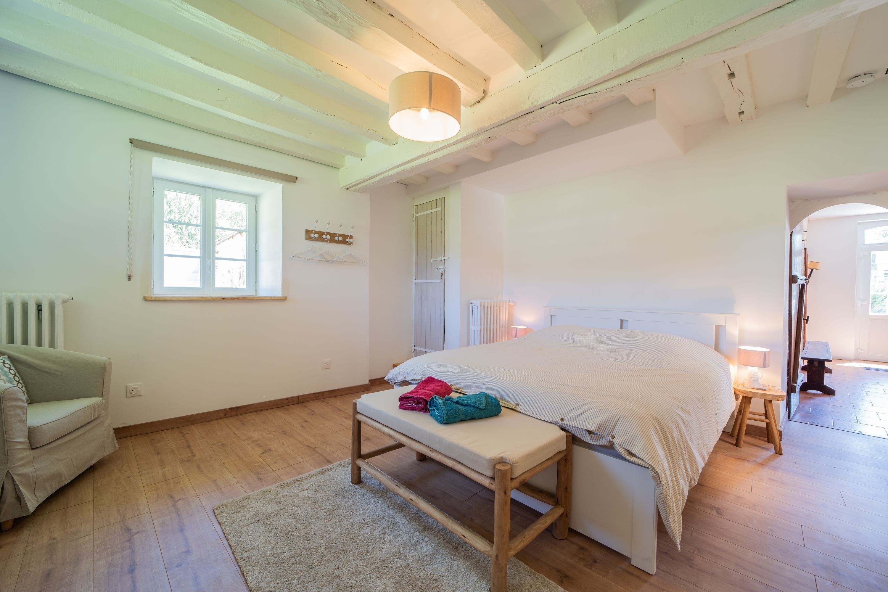 Un dormitorio con cama, banca, silla, ventana y radiador.