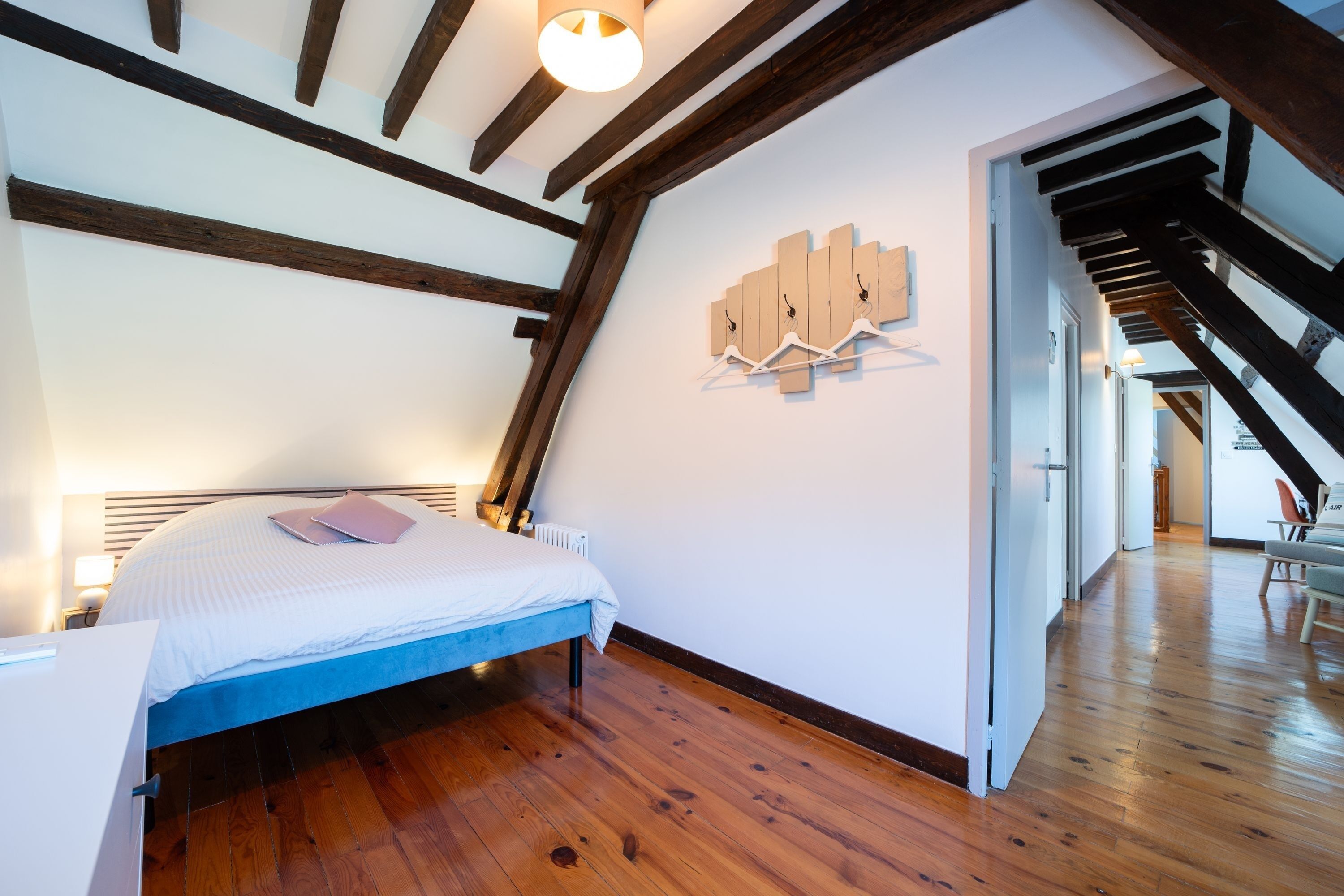 Une chambre à coucher avec un lit, des poutres en bois, un porte-manteau fixé au mur et un couloir menant à une autre pièce.