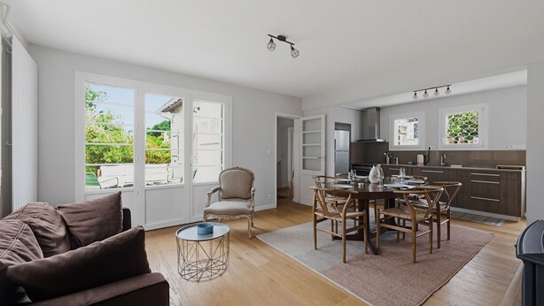 A spacious living area with a sofa, dining table, and chairs, a kitchen with modern appliances, and large windows offering a view of greenery. - Villa ETXEA - À 5 min du port de la Hume (Gujan-Mestras)