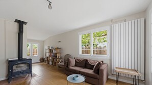A living room with a wooden floor, a brown sofa, a round coffee table, a black wood-burning stove, and a window with a view of greenery. - Villa ETXEA - À 5 min du port de la Hume (Gujan-Mestras)