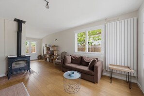 Ein Wohnzimmer mit Holzboden, einem braunen Sofa, einem runden Couchtisch, einem schwarzen Kaminofen und einem Fenster mit Blick ins Grüne.