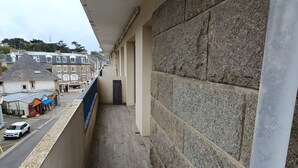 A balcony with a stone wall, a view of a street with shops, and a parked car. - L'as du Val - Avec un accès immédiat sur la plage (Pléneuf-Val-André)