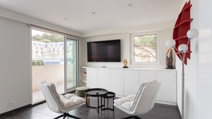 A modern living room with a TV, two white chairs, a small table, and a balcony with a view. - L'as du Val - Avec un accès immédiat sur la plage (Pléneuf-Val-André)