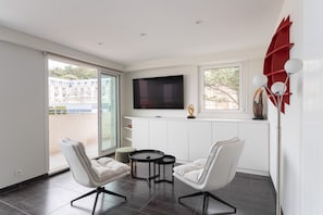 Ein modernes Wohnzimmer mit Fernseher, zwei weißen Sesseln, einem kleinen Tisch und einem Balkon mit Blick.
