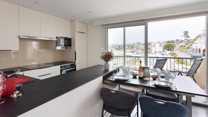 A modern kitchen with a large island, a dining area with chairs, and a view of the city through large windows. - L'as du Val - Avec un accès immédiat sur la plage (Pléneuf-Val-André)