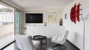 A modern living room with a flat-screen TV, two white armchairs, a small round table, and a red abstract wall decoration. - L'as du Val - Avec un accès immédiat sur la plage (Pléneuf-Val-André)