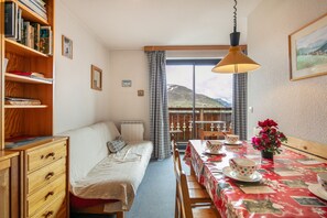 A room with a bed, a wooden dresser, a dining table with a red tablecloth, a potted plant, and a view of a mountain through a window. - La Fontaine du Corbeau et Grangettes (Huez)