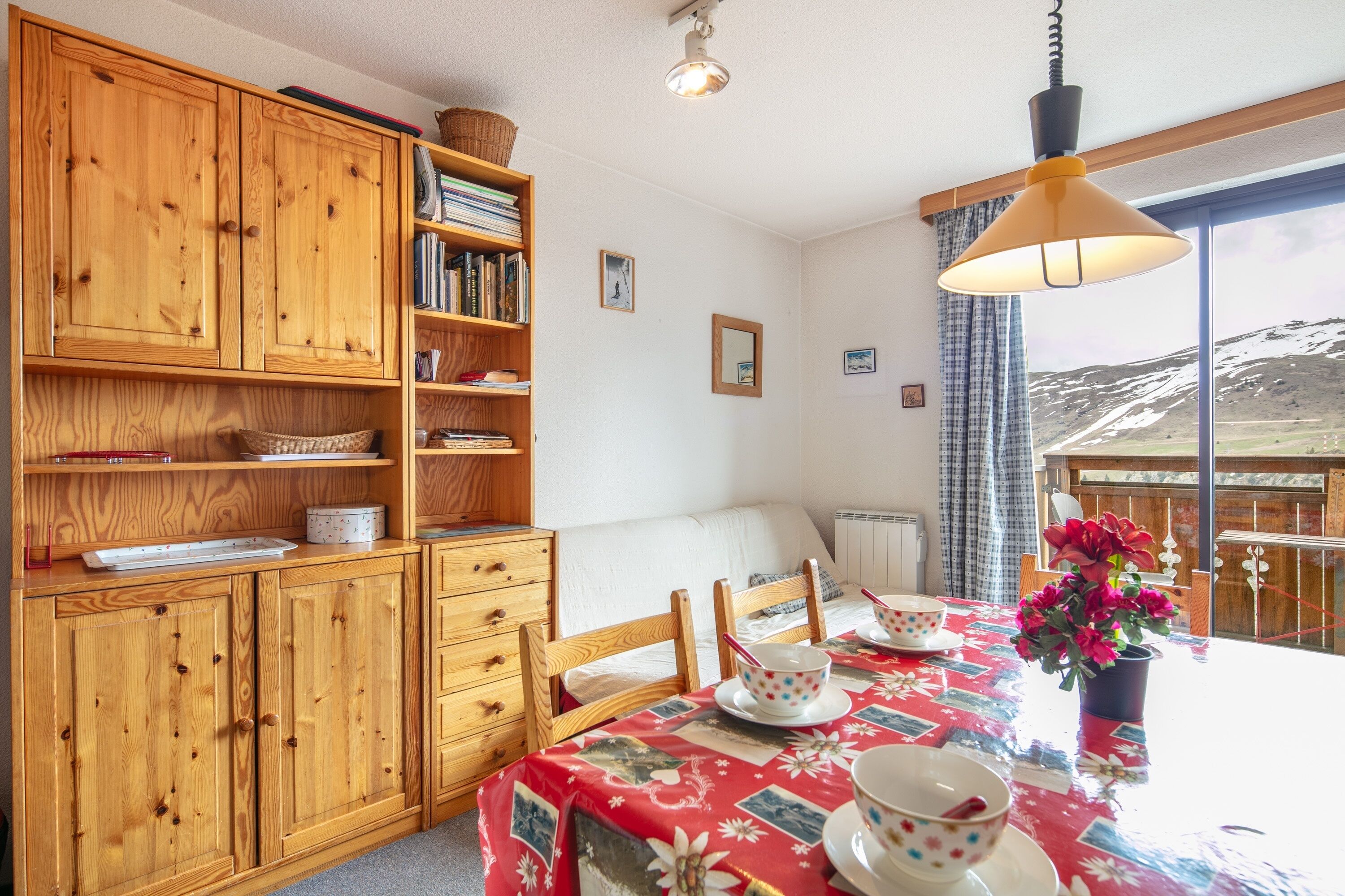 A room with wooden cabinets, a dining table with a floral tablecloth, chairs, and a view of a snowy landscape through a window.