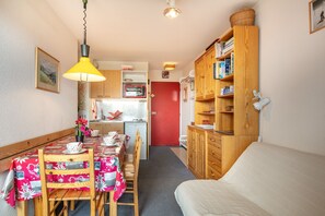 A compact room with a dining table, chairs, a bed, a kitchenette, and a bookshelf. - La Fontaine du Corbeau et Grangettes (Huez)