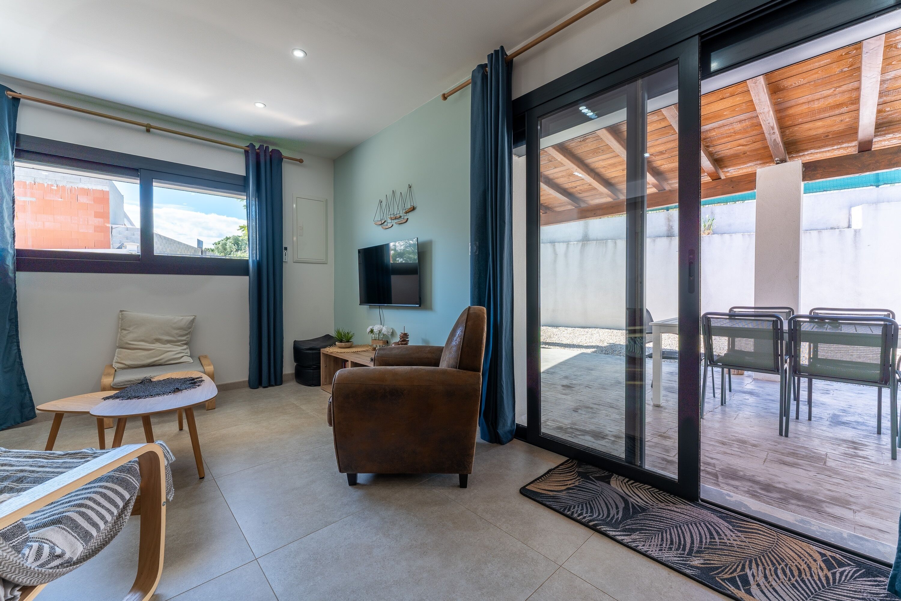 Un salon moderne avec un canapé, un fauteuil et une petite table. Un téléviseur est fixé au mur, et une porte-fenêtre coulissante donne accès à un espace extérieur aménagé avec des chaises.