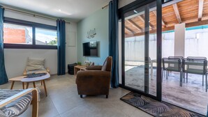 Een moderne woonkamer met een bank, fauteuil en een salontafel. Er is een televisie aan de muur bevestigd en een schuifpui die uitkomt op een buitenruimte met stoelen.
