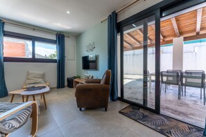 Ein modernes Wohnzimmer mit einer Couch, einem Sessel und einem kleinen Tisch. Ein Fernseher ist an der Wand montiert, eine Glastür führt zu einem Außenbereich mit Stühlen.