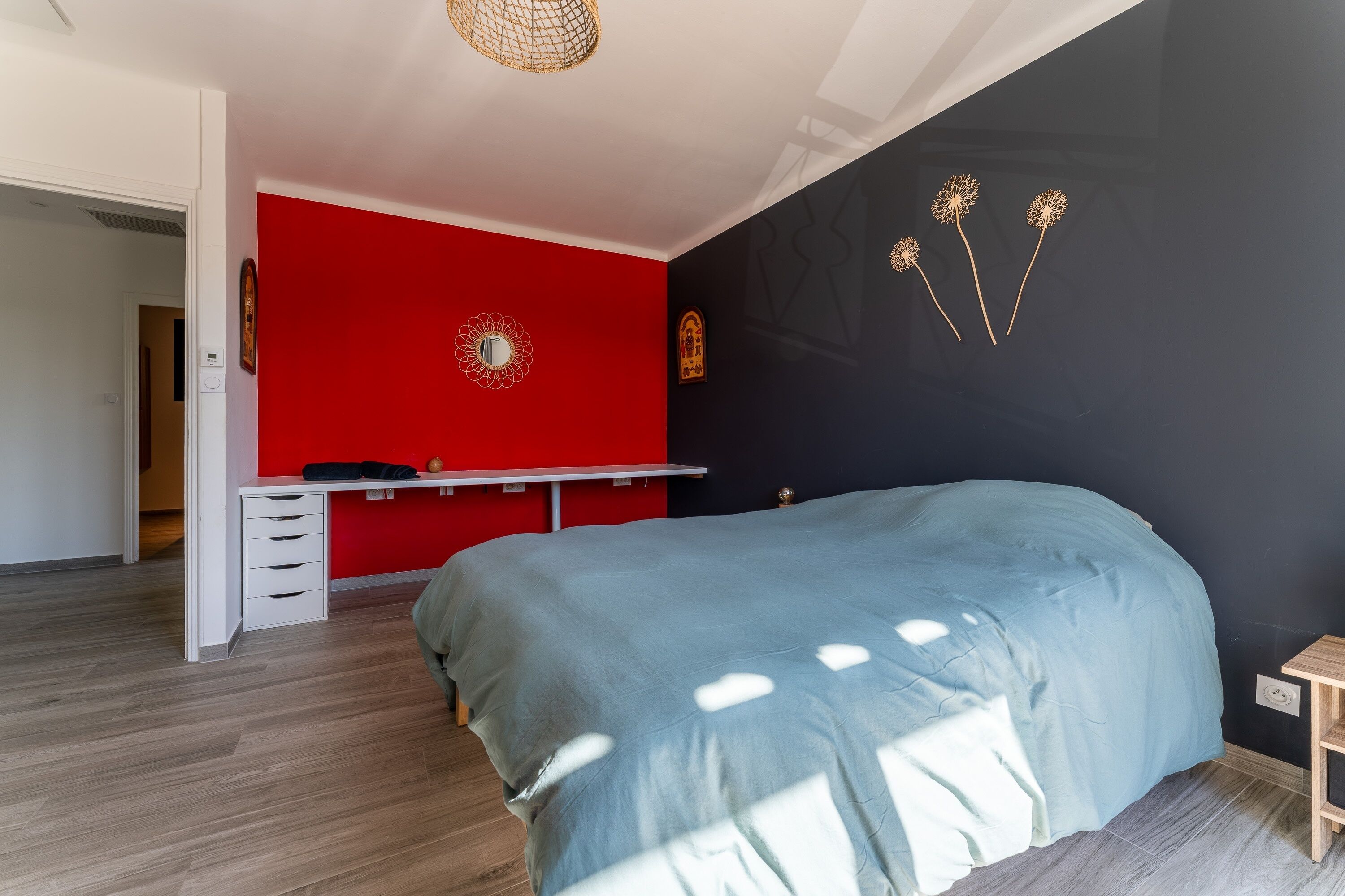 Une chambre à coucher avec un lit, un bureau et une chaise. Un mur est peint en rouge et orné d’un objet décoratif, tandis qu’un autre mur est noir et présente une œuvre d’art murale.