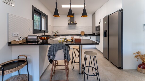 Een moderne keuken met een centraal eiland, roestvrijstalen apparatuur, hanglampen en uitzicht op de buitenlucht.