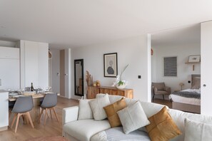 A modern living room with a sofa, a dining area with chairs, a wooden cabinet, and a bed in the background. - Le Madison - À moins d'1 km de la plage (Pléneuf-Val-André)