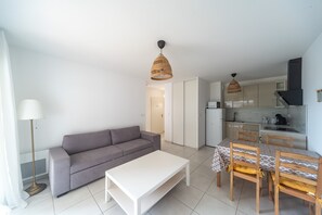 A modern living room with a sofa, a white coffee table, and a dining area with a table and chairs. There is a kitchen area with cabinets and a stove. - Le Lavandou Ensoleillé - Bel appt avec terrasse (Le Lavandou)