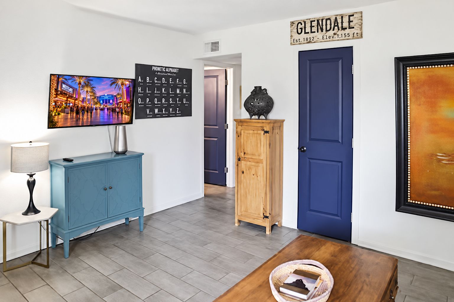 Un couloir avec une porte bleue, une armoire bleue, un tableau encadré représentant une vue de la ville, une affiche de l’alphabet phonétique et une pancarte sur laquelle on peut lire « GLENDALE ».