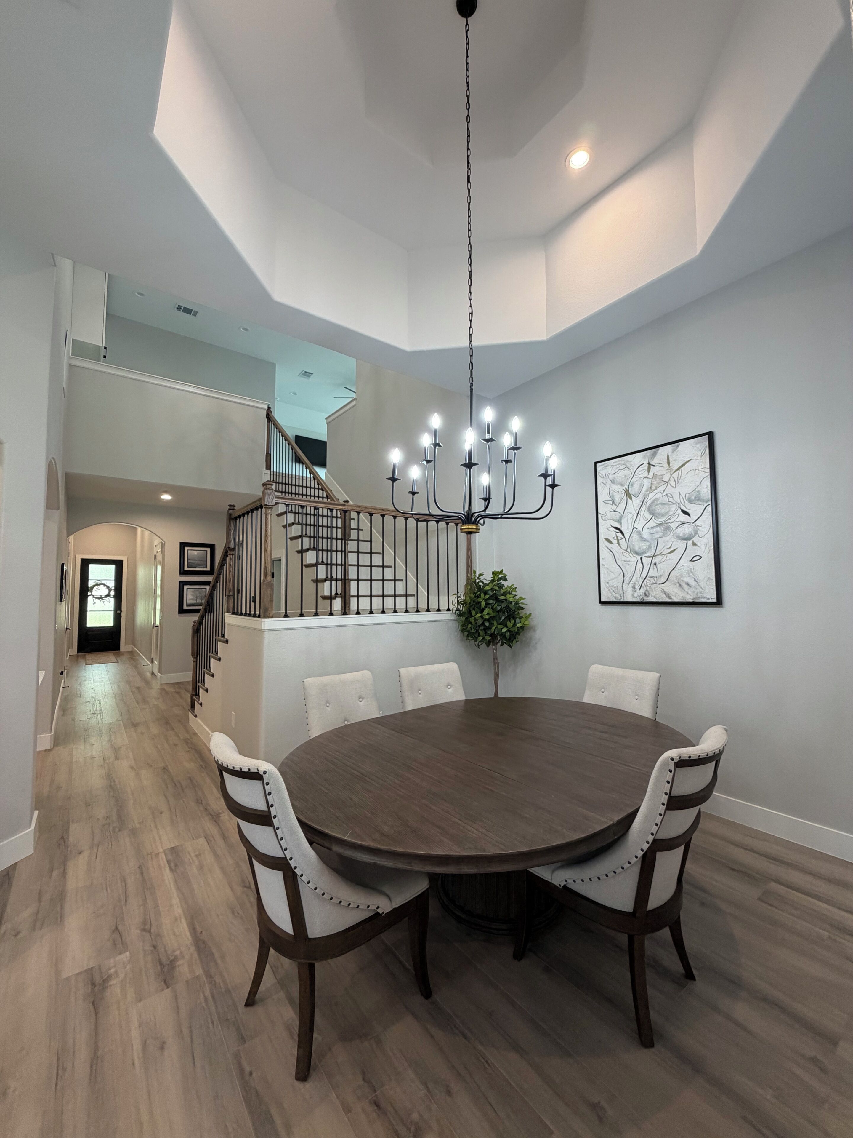 A dining area with a round table, six chairs, a chandelier, a staircase, and a framed artwork on the wall.
