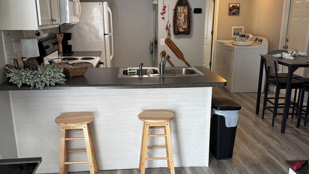 A modern kitchen with white cabinets, a central island with stools, a washing machine, and a dining area with a table and chairs. - Lovely 2-bedroom mobile home with AC in Corbin. Only .4 miles from Laurel Lake. (Corbin)