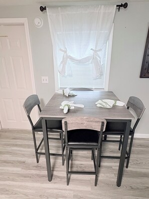 A dining table with four chairs, a window with blinds, a door, and a wall-mounted light switch.