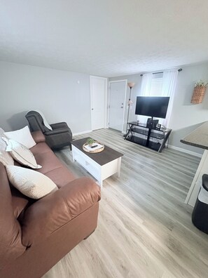 A living room with a brown sofa, a white coffee table, a TV, and a dining area with a table and chairs.