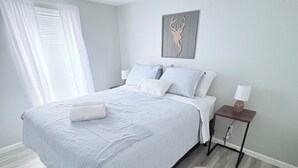 A bedroom with a bed, a bedside table, a lamp, a decorative wall piece, and a window with curtains. - Lovely 2-bedroom mobile home with AC in Corbin. Only .4 miles from Laurel Lake. (Corbin)