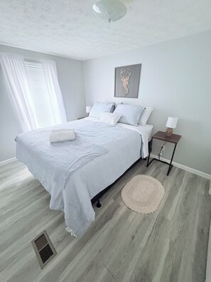 A bedroom with a bed, a bedside table, a lamp, a decorative wall piece, and a window with curtains.