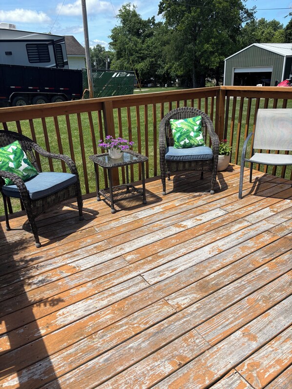 Une terrasse en bois avec des chaises en osier et une petite table, un abri de jardin vert et, au fond, un camping-car blanc.