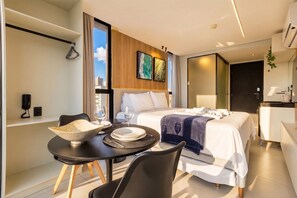 Ein modernes Hotelzimmer mit Bett, Esstisch mit Stühlen, einer kleinen Küche und einem großen Fenster.
