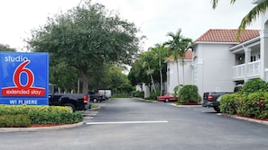 Ein Studio 6 Hotel für längeren Aufenthalt mit Parkplatz, Bäumen und einem Gebäude mit rotem Ziegeldach.