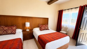 Habitación de hotel con dos camas, una mesita de noche con lámpara, una silla y vistas al exterior a través de una ventana con cortinas.