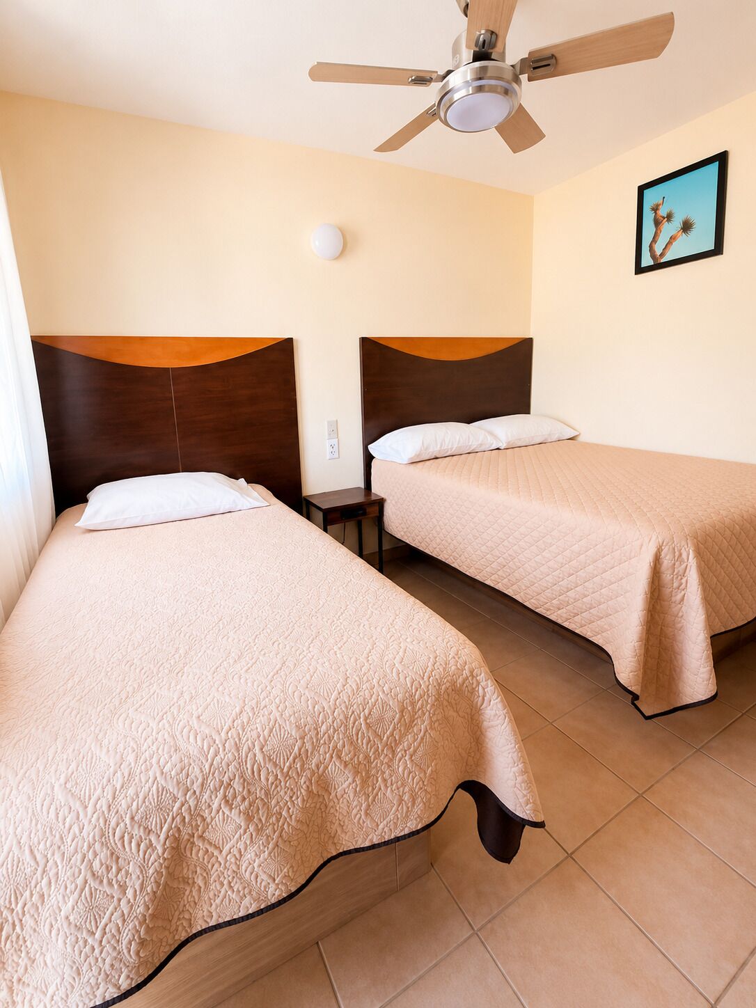 A hotel room with two beds, a ceiling fan, a nightstand, a wall-mounted light, and a framed picture.