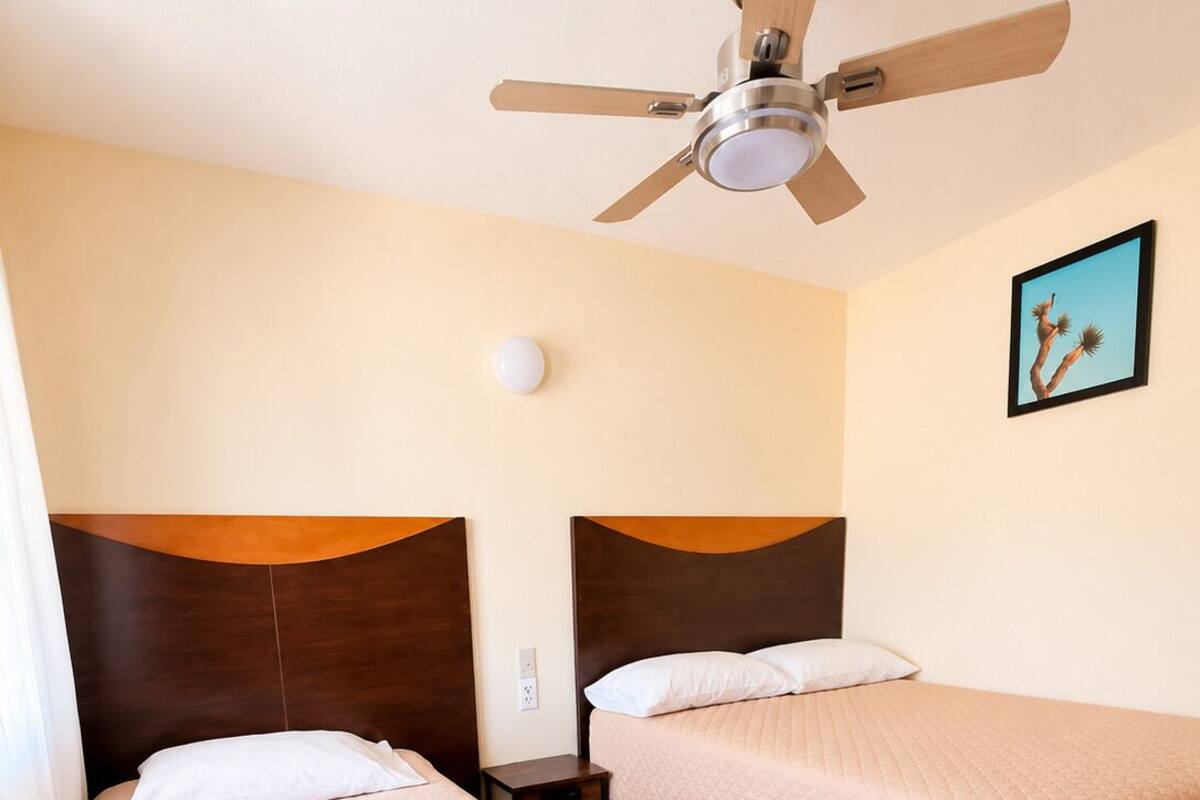 A hotel room with two beds, a ceiling fan, a nightstand, a wall-mounted light, and a framed picture.