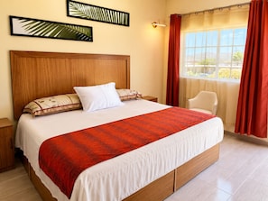 A hotel room with a large bed, a wooden headboard, a nightstand, a chair, and a window with curtains. - Hotel Las Misiones (El Rosario)