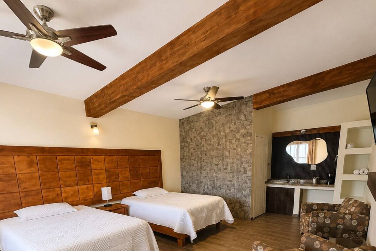 A hotel room with two beds, a wooden ceiling, a stone accent wall, a small desk, a chair, and a ceiling fan.