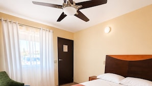 Habitación de hotel con cama, ventilador de techo, una silla, una puerta y una ventana con cortinas.