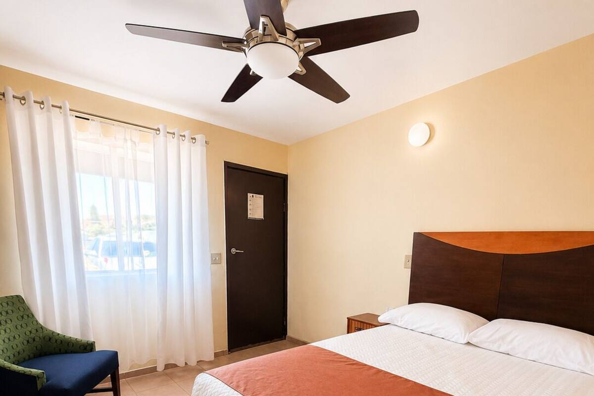 A hotel room with a bed, a ceiling fan, a chair, a door, and a window with curtains.