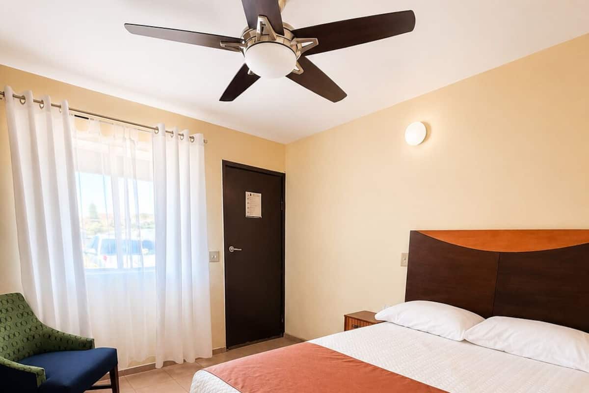 A hotel room with a bed, a ceiling fan, a chair, a door, and a window with curtains.
