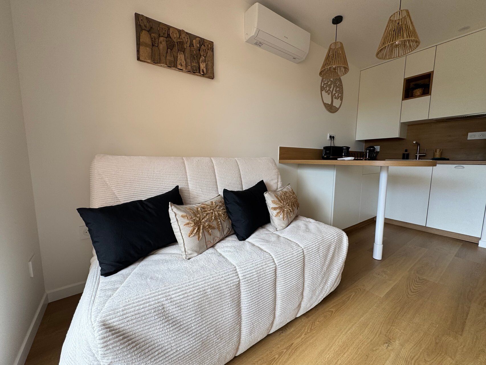 A modern living room with a white sofa, black and beige cushions, a kitchen area with white cabinets, and a hanging light fixture.