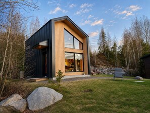 A modern two-story house with a large window, a wooden exterior, and a stone wall in the background. - Lakeside Cabin Retreat with Beach, Kayaks, Fire Pit & Forest Trails (Sainte-Marguerite-du-Lac-Masson)