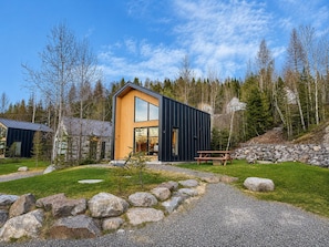En moderne hytte med et stort glasvindue, omgivet af et naturligt landskab med træer og en grussti.