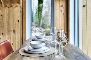 A dining table set with bowls, plates, and glasses, with a view of trees and rocks through a window. - Lakeside Cabin Retreat with Beach, Kayaks, Fire Pit & Forest Trails (Sainte-Marguerite-du-Lac-Masson)