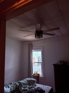 Ein Schlafzimmer mit Deckenventilator, einem Fenster mit Blick auf einen Zaun, einem Bett mit grüner Decke und einem Schminktisch.