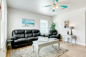 Ein Wohnzimmer mit einem schwarzen Ledersofa, einem weißen Couchtisch, einem Deckenventilator, einem Gemälde mit Strandmotiv und einer kleinen Pflanze.