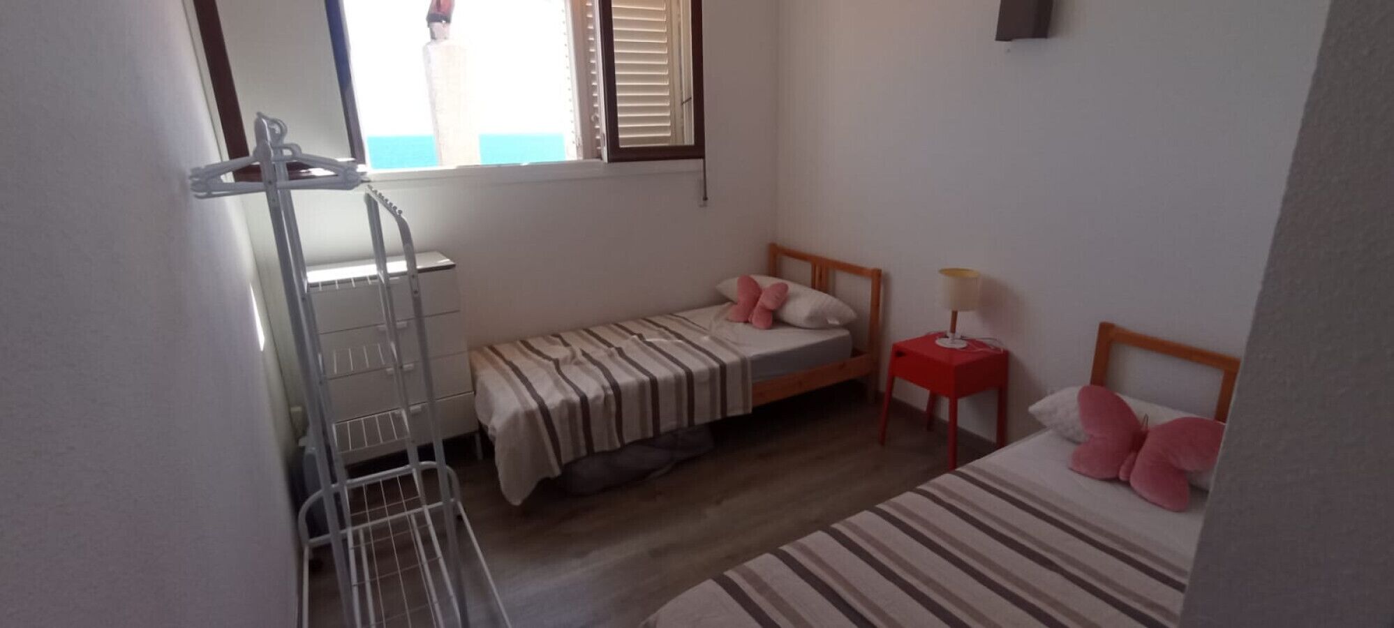 Un dormitorio con dos camas, una ventana con persianas, una mesita con lámpara y un perchero de metal.