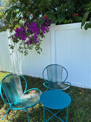 To turkis metalstole og et lille rundt bord i en have med hvid hegn og lilla blomster.