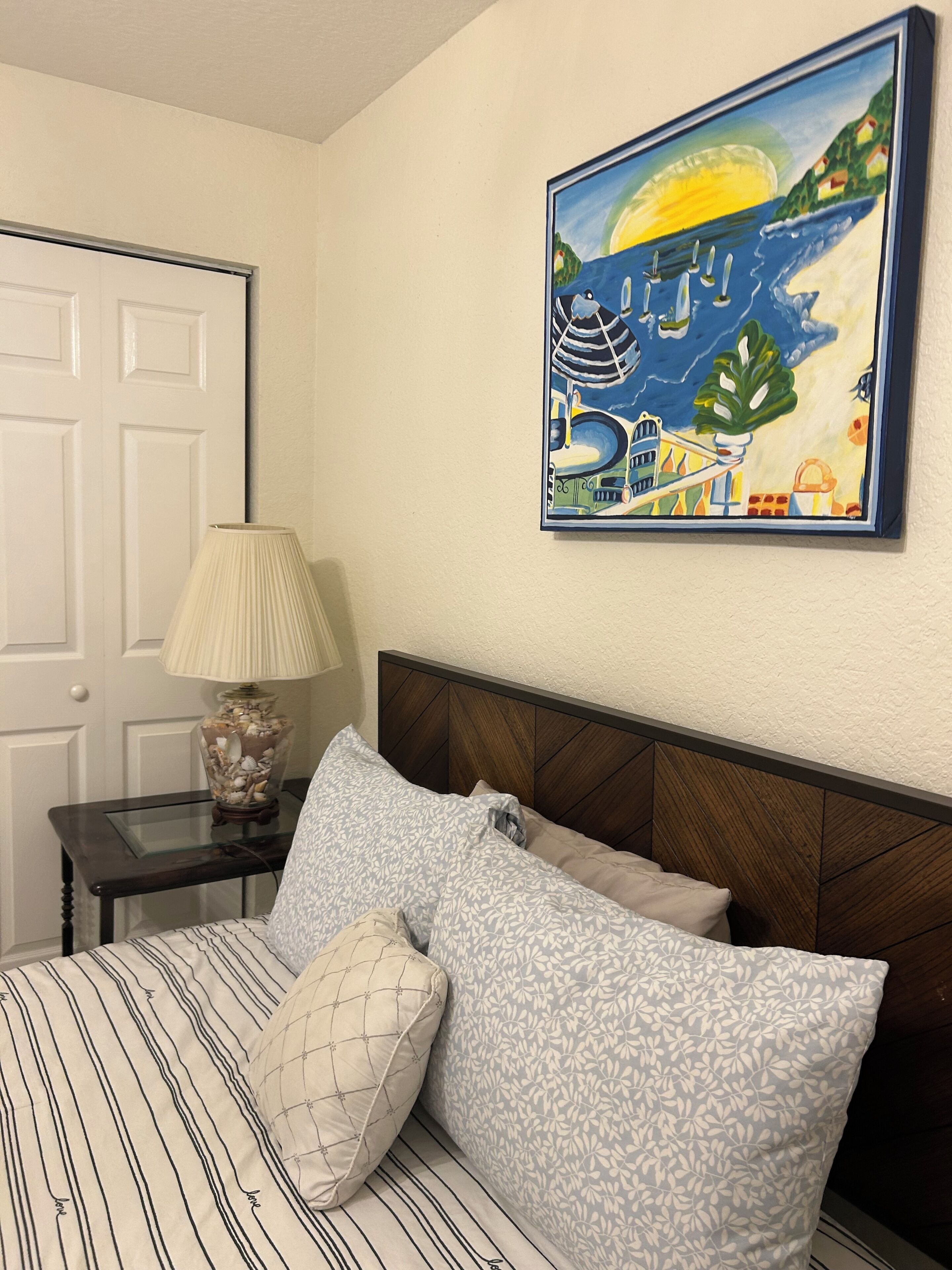 Een slaapkamer met een bed, een nachtkastje met een lamp, een aan de muur hangend schilderij en een deur.