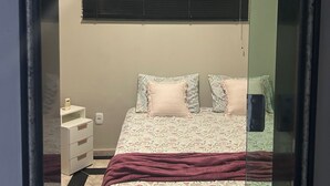 Een slaapkamer met een bed, nachtkastje, spiegel en een raam met jaloezieën.