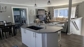 A modern kitchen with a central island, a dining area with chairs, a sofa, and a staircase leading to a balcony. - Indoor Pool! Hot Tub! Sauna! at the Blue Haven! (Trent Lakes)