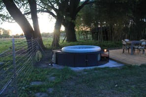 A hot tub with a wooden deck, surrounded by a fenced area with trees and a table with chairs.